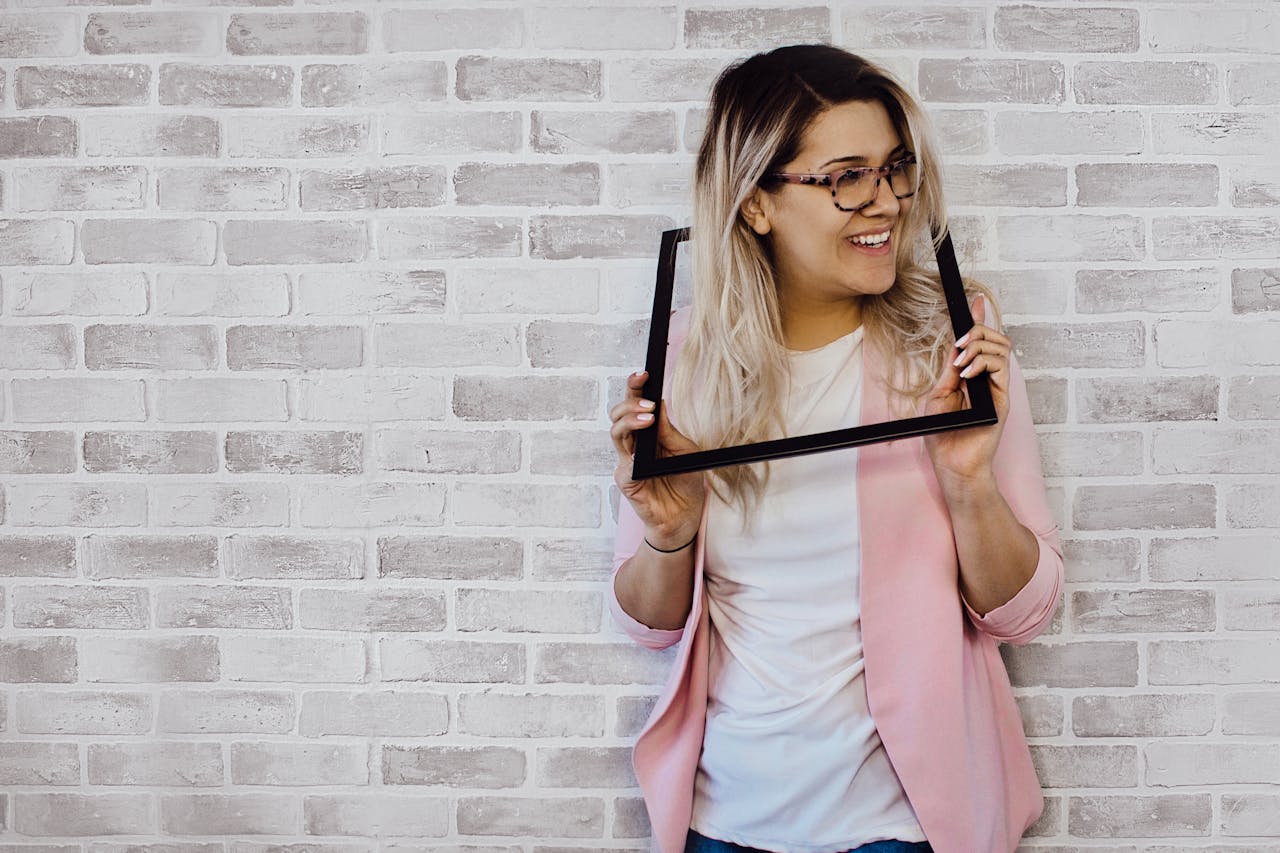 Cheerful woman with blonde hair posing creatively with a picture frame in a stylish indoor setting.